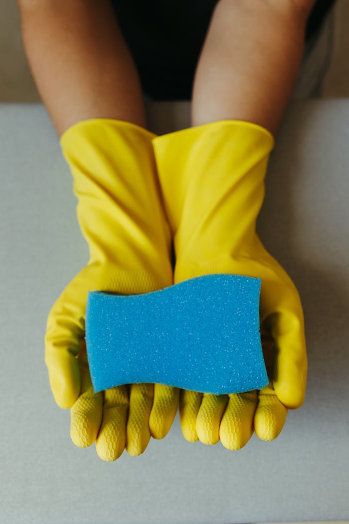 services-04 Close-up of yellow gloves holding a blue cleaning sponge, ideal for home cleaning themes.