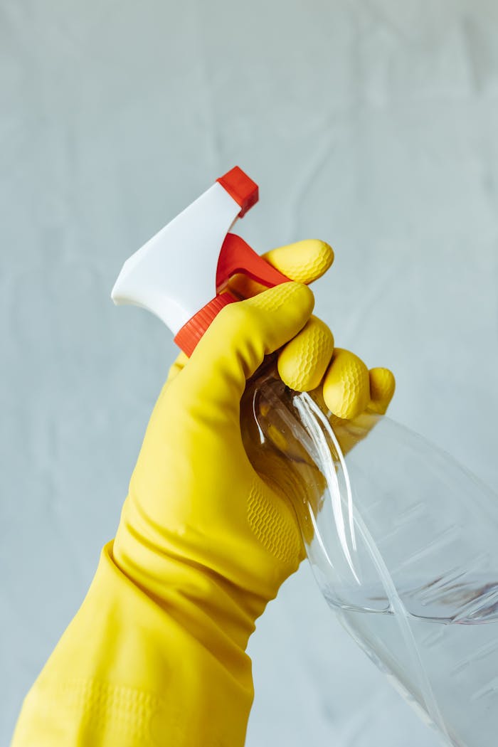 hero-img A hand wearing a yellow rubber glove holds a red and white spray bottle against a neutral background.